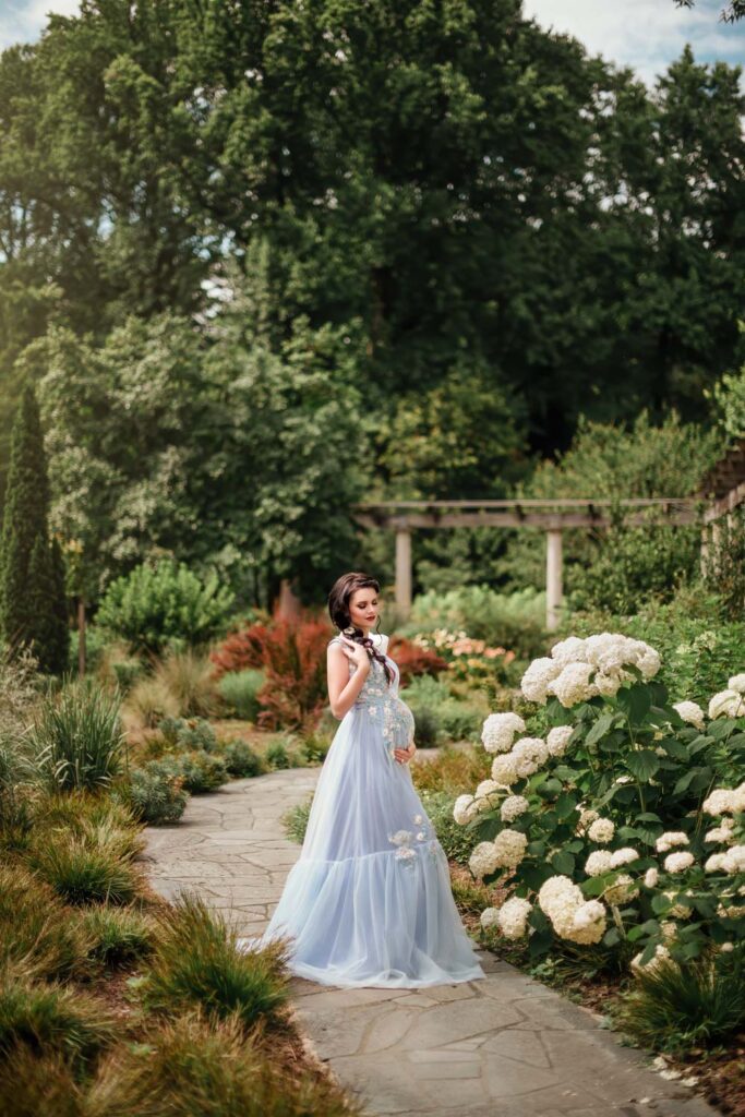 pregnant woman in a pale blue embroidered gown on a curved garden path bordered by white hydrangeas.