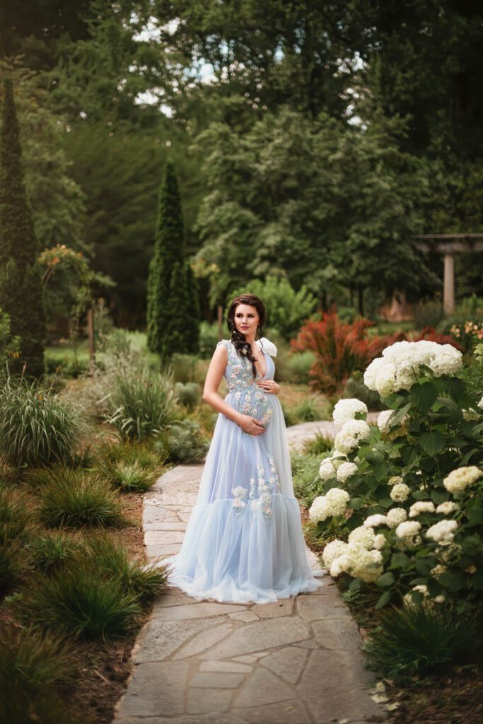 Mom-to-be in a pale blue embroidered gown cradles her belly on a garden path beside clusters of white hydrangeas.