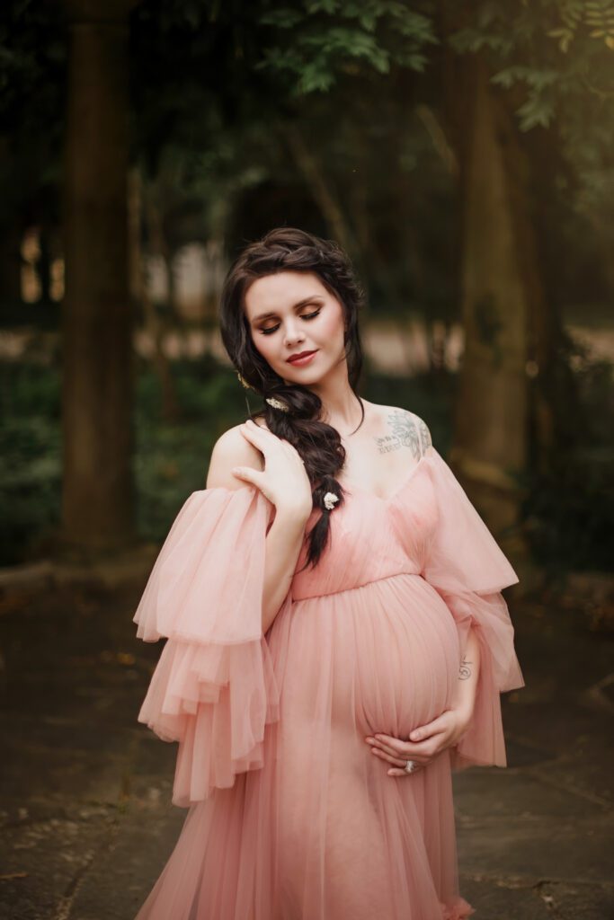 Expecting mother in a blush ruffled gown cradling her belly among trees at Cator Woolford Gardens in Atlanta.