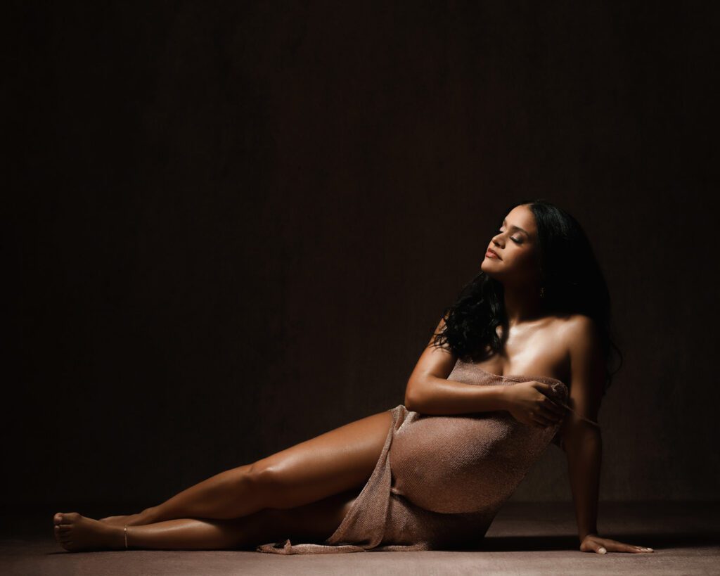 Pregnant woman in a bronze gown laying on studio floor propped up on one arm cradling her belly during Marietta maternity photos.