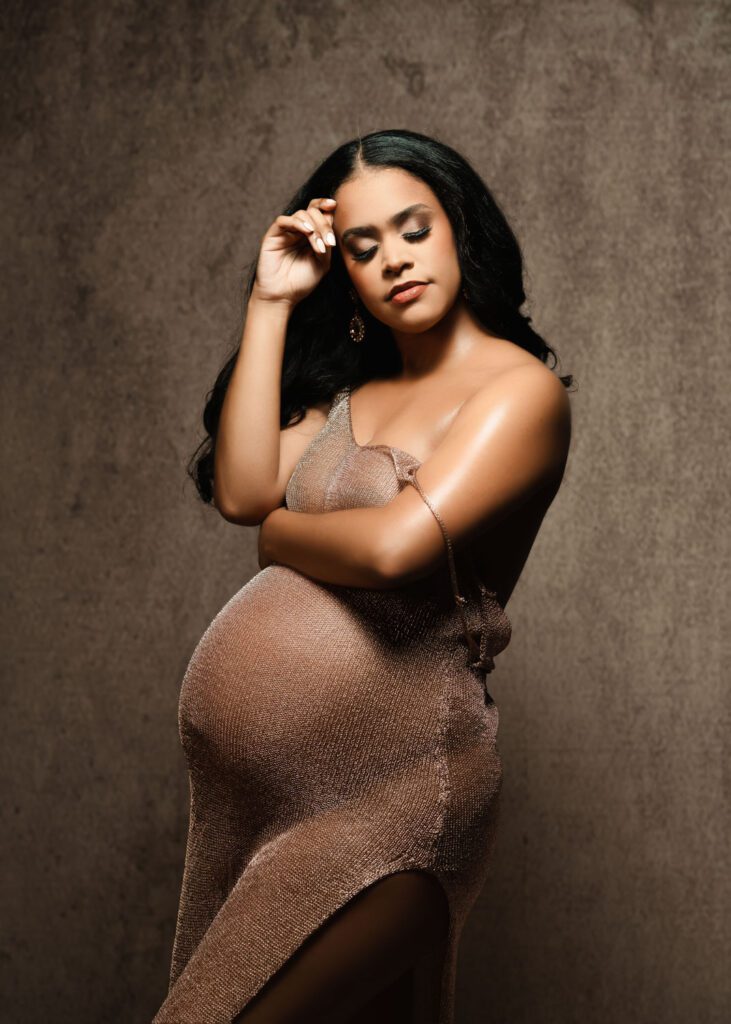 Pregnant black woman in a fitted bronze gown with one arm raised and the other resting across her belly.