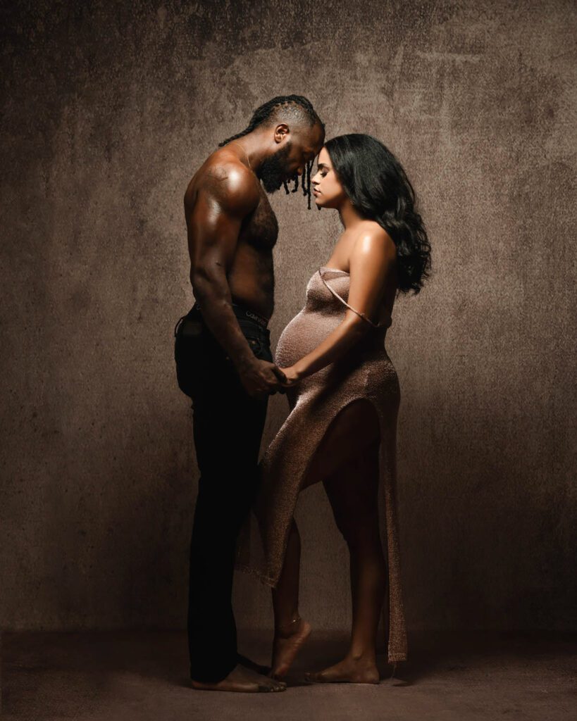 Black couple holding hands touching foreheads and eyes closed standing in front of brown textured backdrop with dramatic lighting. 
