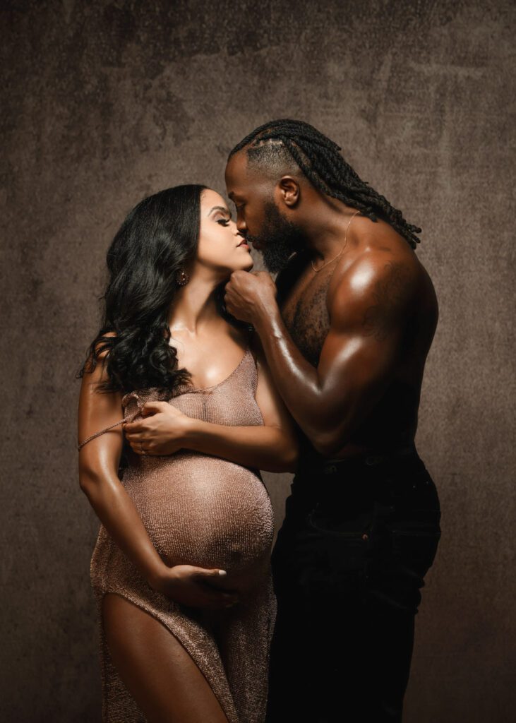 Muscular dad with oiled skin touches his pregnant wife's chin posed in front of brown textured backdrop in studio maternity photoshoot.