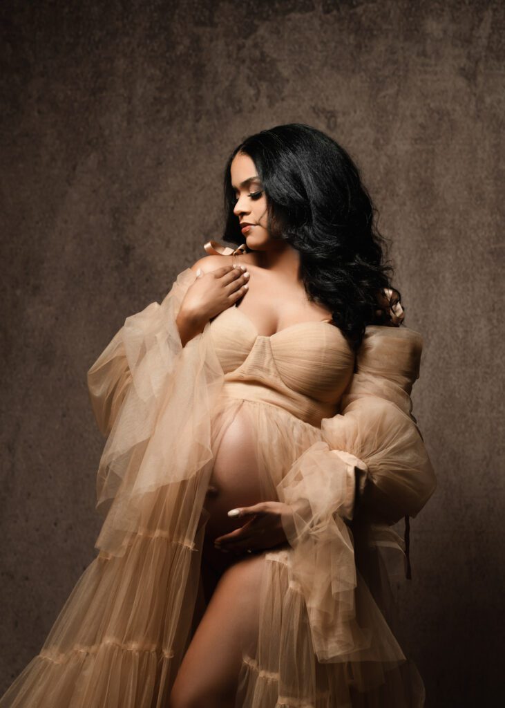 Pregnant woman in a beige tulle gown looking down with one hand resting near her bare belly.