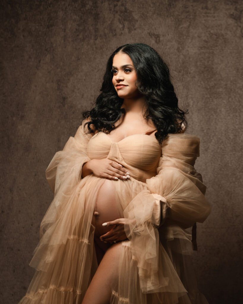 Pregnant woman in a beige tulle maternity gown cradling her bare belly against a textured brown studio backdrop.