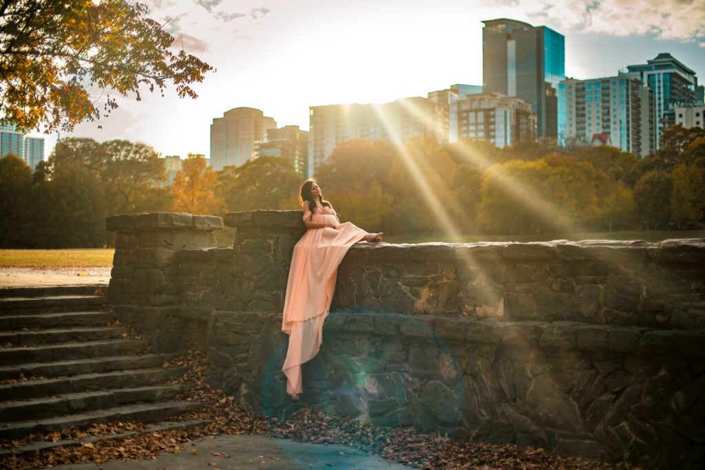 Expecting mother in a soft peach gown sits along a stone wall as golden sunlight streams over the Atlanta city skyline.