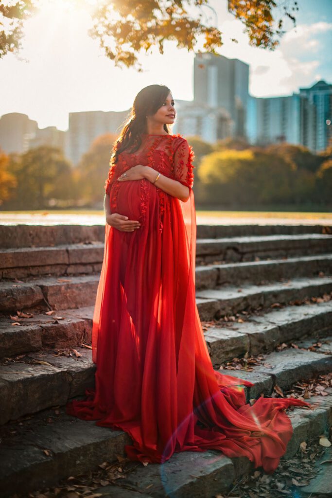 pregnant mom in a dramatic red gown on stone steps with warm sunlight and the Atlanta skyline behind her