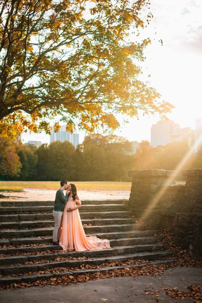Couple shares a kiss on stone steps while the expecting mother cradles her belly in a long peach gown with a flowing train