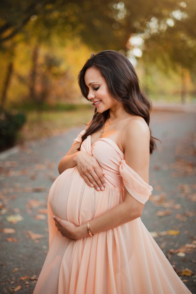 smiling pregnant woman wearing a sweetheart peach maternity gown with a path and trees of Oak Hill in the background