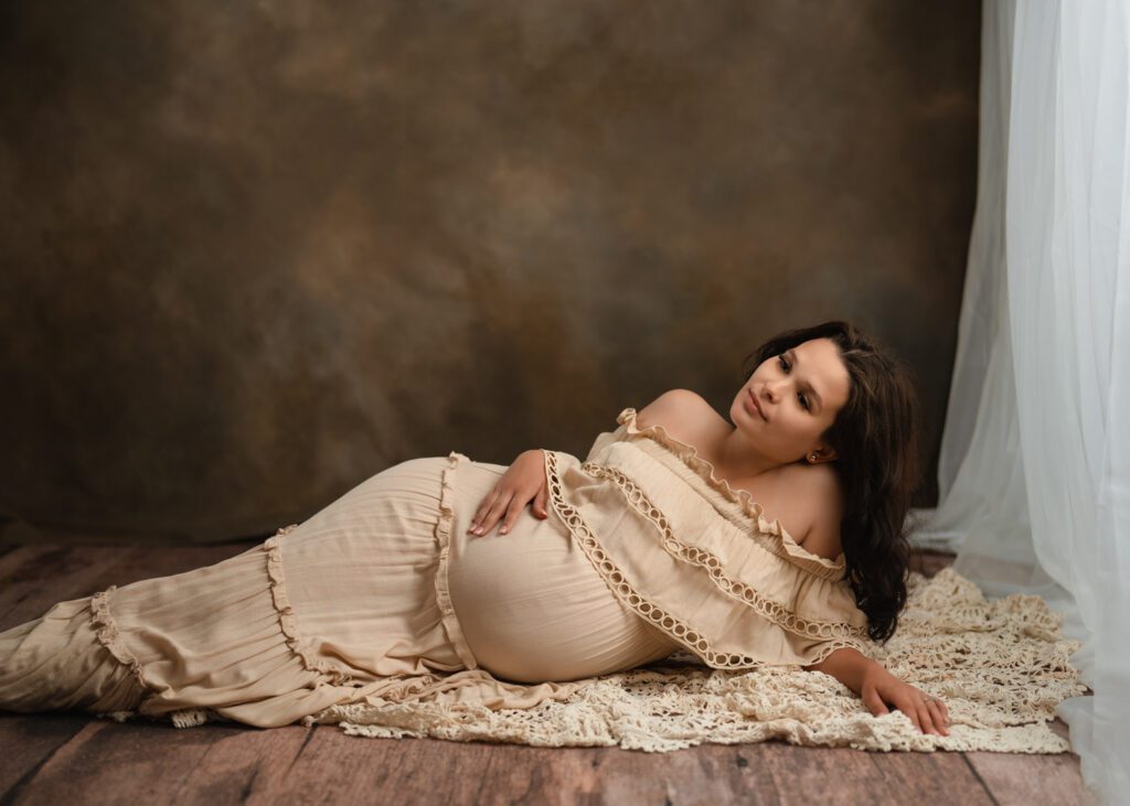 Pregnant expecting mom laying on hardwood floor in tan gown during maternity photoshoot in studio.