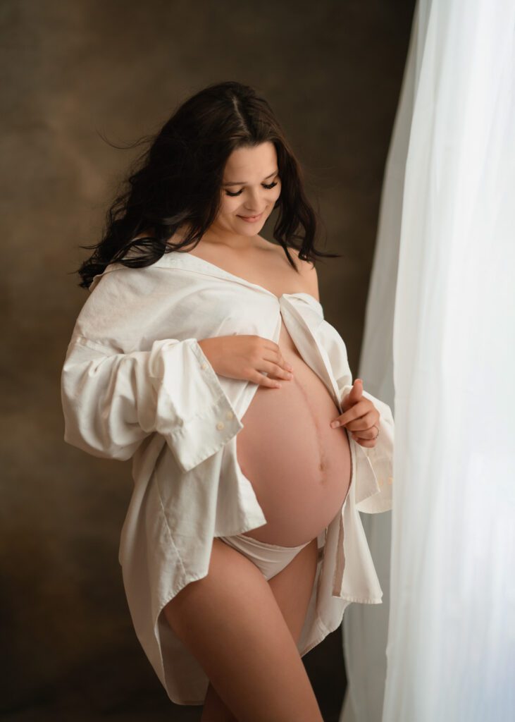 Expecting mother playing with her belly bump awaiting baby's arrival in white  shirt in front of window.