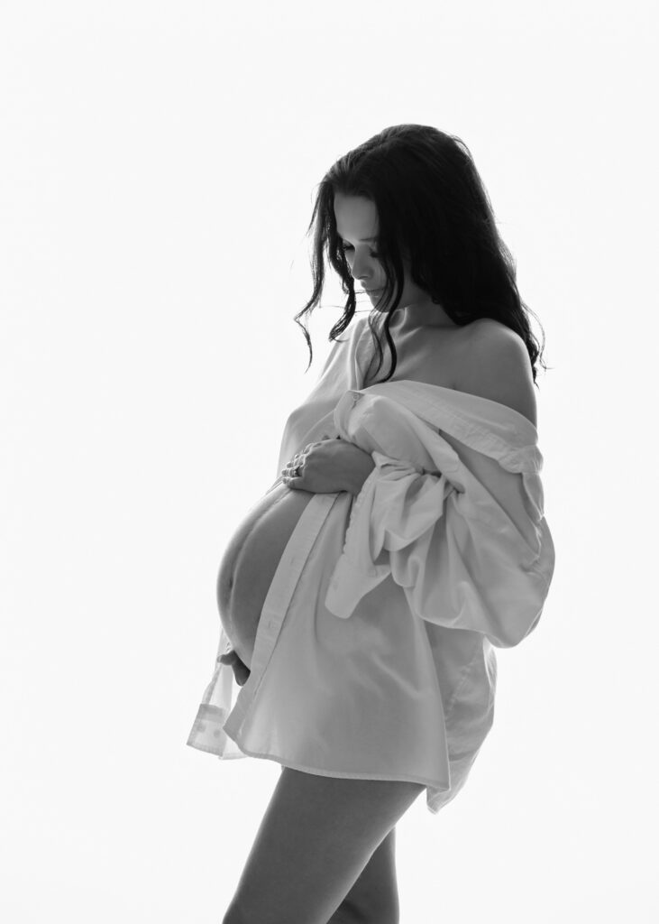 Pregnant woman in a white robe with one hand above her belly against a bright softly lit studio backdrop.