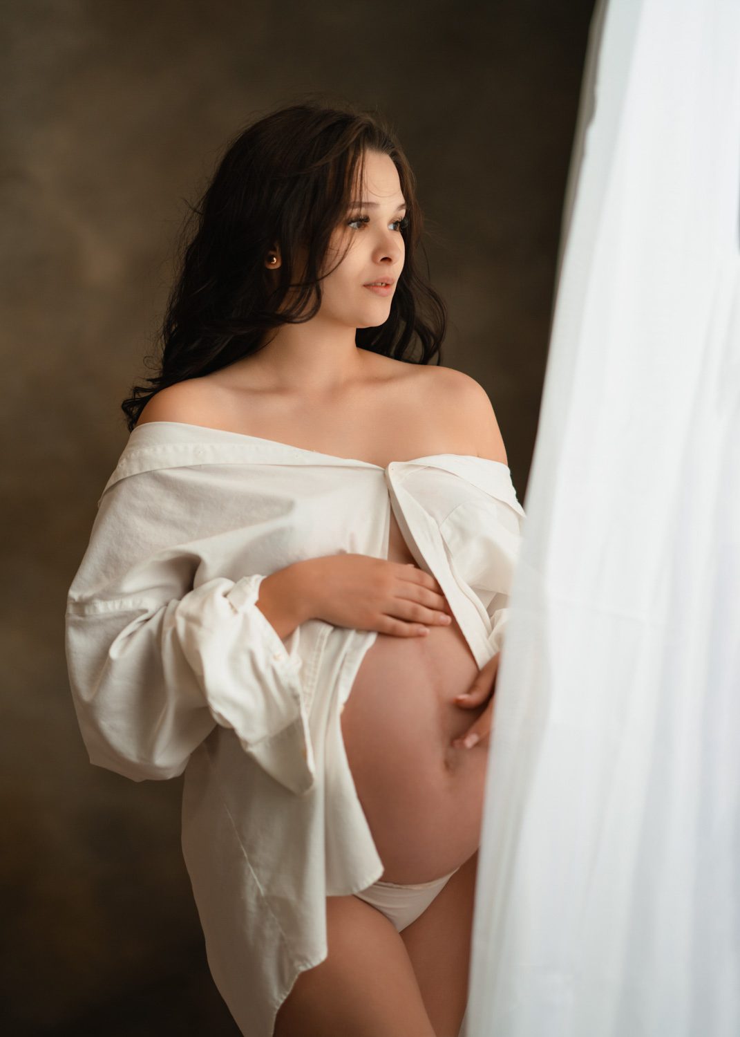 pregnant mom in white mens shirt holding her belly beside a softly lit window photographed by roswell maternity photographer