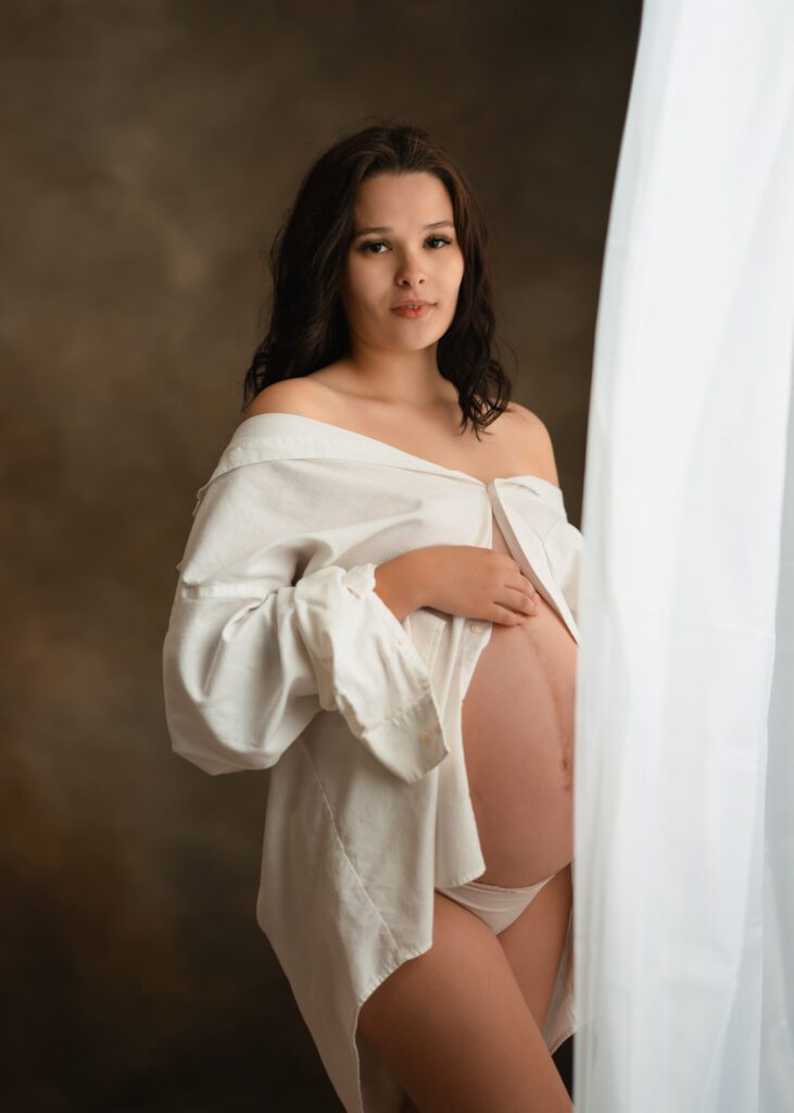 Expecting mother in a white shirt cradling her belly beside soft window light for Roswell maternity photos.