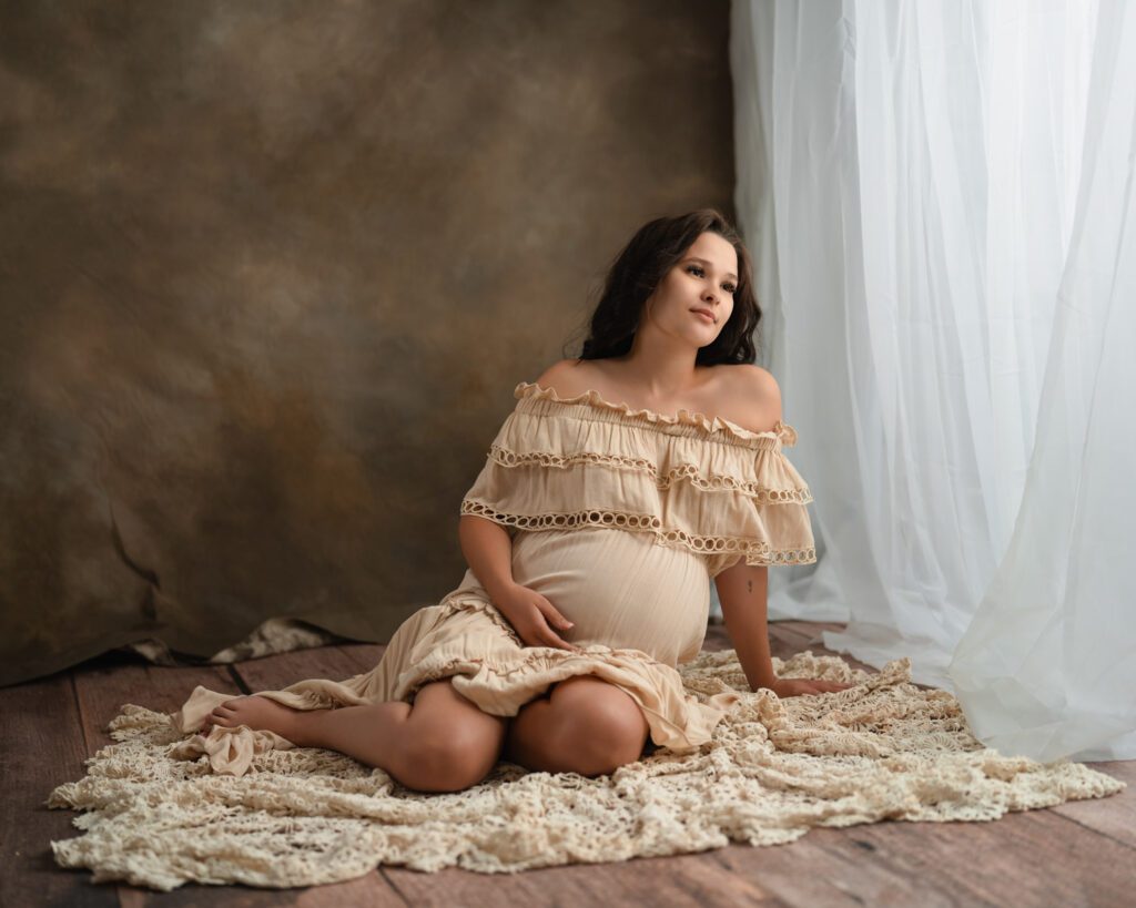 Pregnant woman in a cream off-the-shoulder gown seated on a textured blanket beside a bright window.