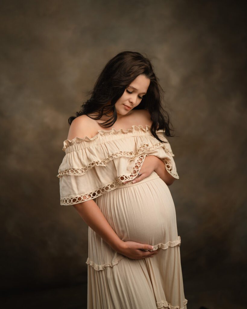 Expecting mother in a cream gown looking down while cradling her belly against a warm studio backdrop.