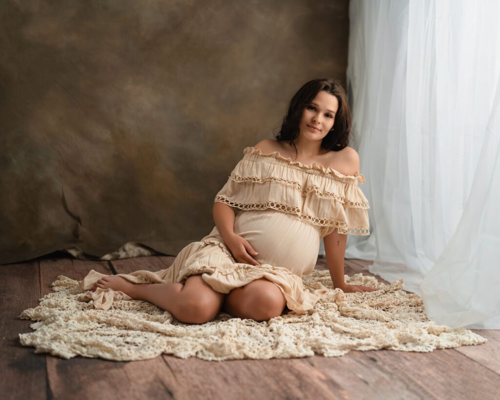 Pregnant woman in a cream ruffled gown sitting on a textured blanket against a warm brown studio backdrop.