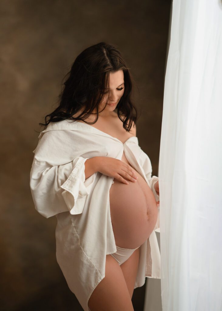 Expecting mother in open white shirt toucing her belly gently beside a softly lit studio window.