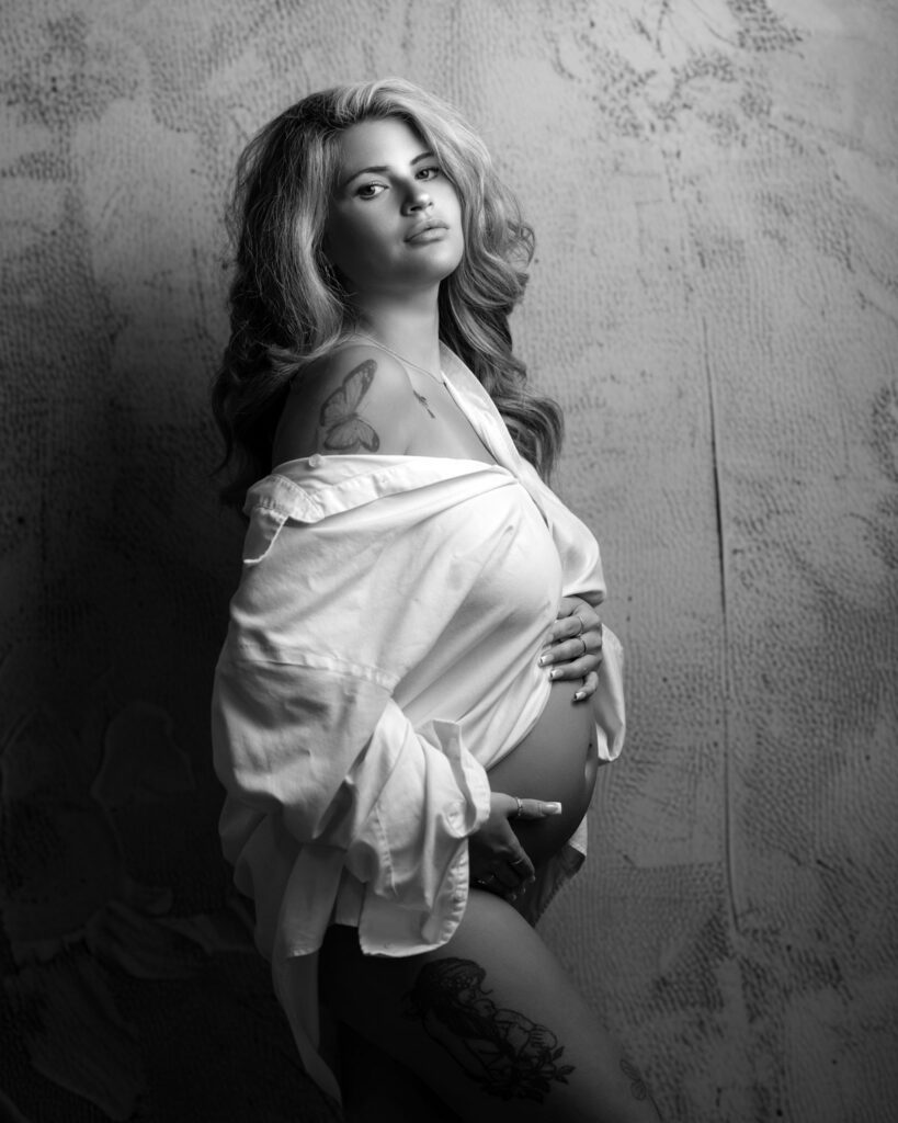Black and white maternity portrait of a pregnant woman in an open white shirt cradling her bare belly against a textured wall.