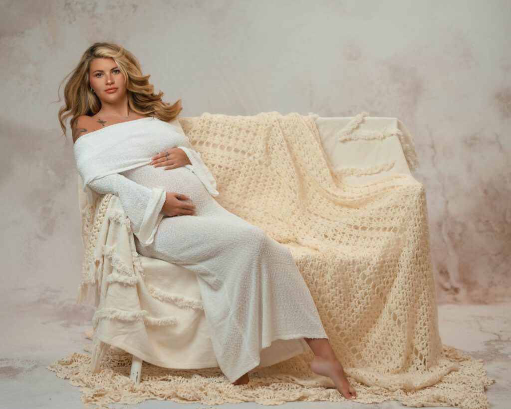 Pregnant woman in a white gown seated on a draped chair, cradling her belly against a soft neutral studio backdrop.