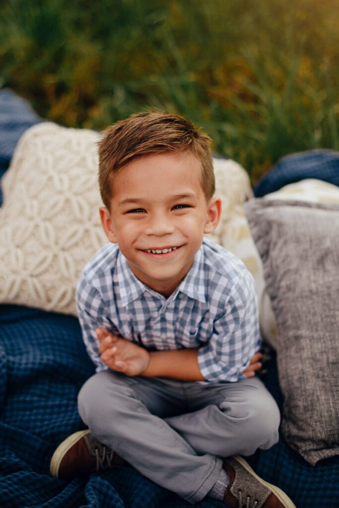 smiling young boy in jeans sitting criss cross on blue blanket with cream pattern pillows.