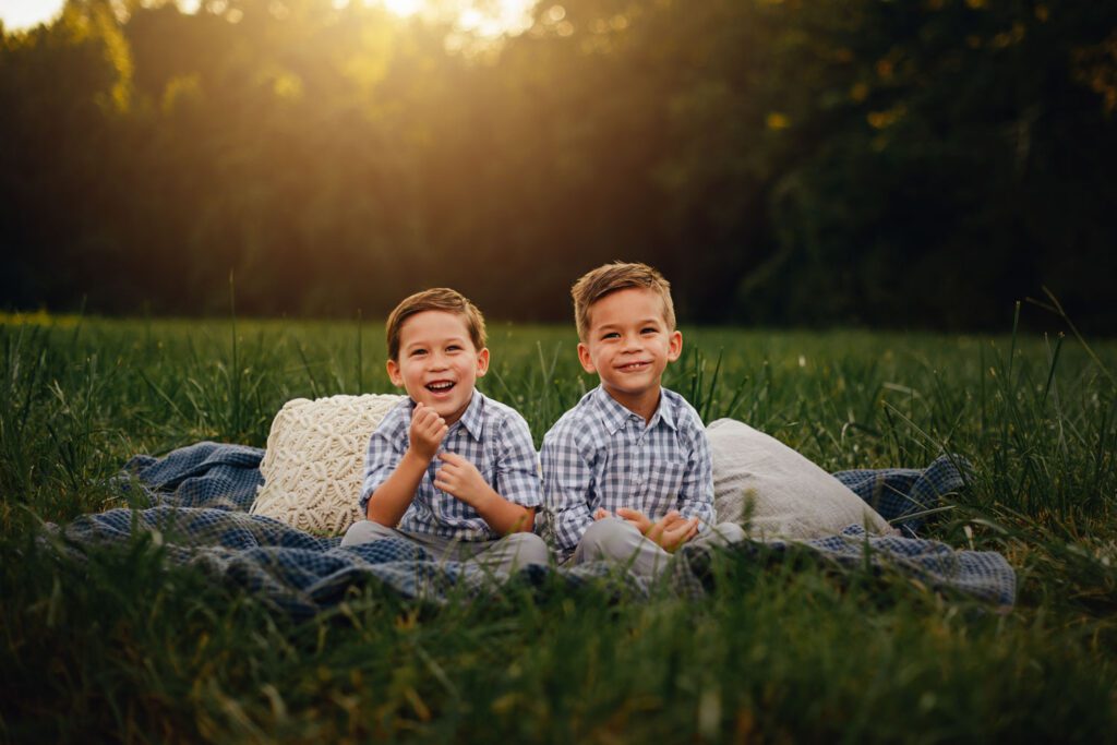 twin brothers in matching plaid shirts sitting on a blanket in grassy meadow at sunset