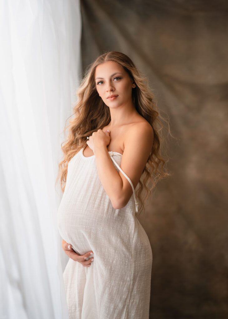 Expecting mother wrapped in white fabric beside a bright window during an Alpharetta maternity photography session.