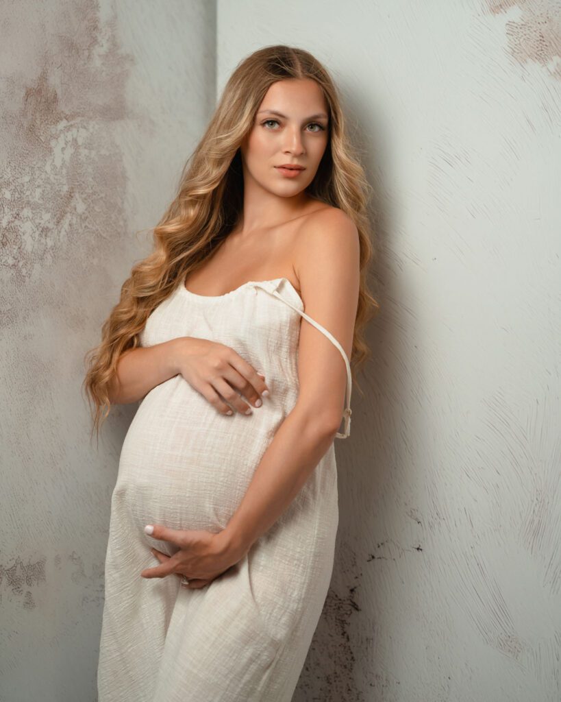 pregnant mom looking at camera in white dress with both hands gently resting around her belly.