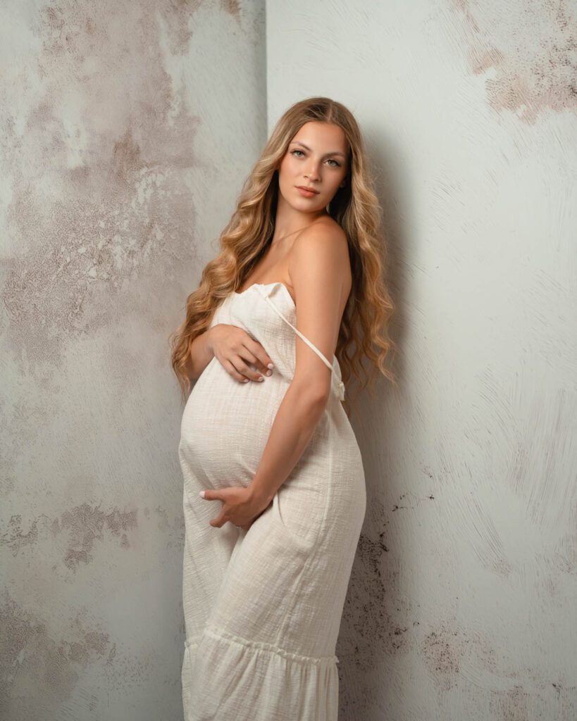 Expecting mother in a cream textured gown cradling her belly against a light textured wall during Alpharetta maternity photos.