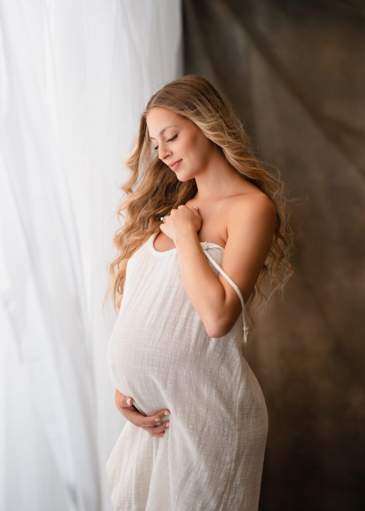 Expecting mother in dress looks down at her belly near window photographed by Alpharetta maternity photographer