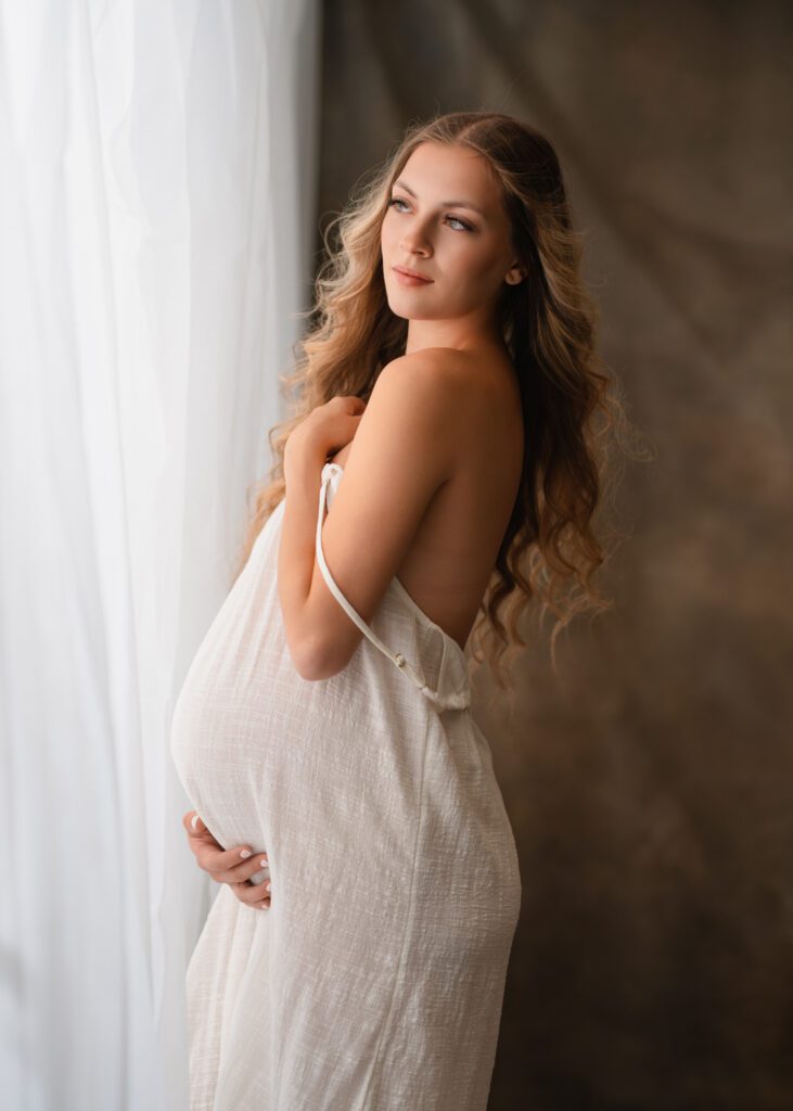 Expecting mother in a white linen dress standing beside a bright window against a warm brown studio backdrop.
