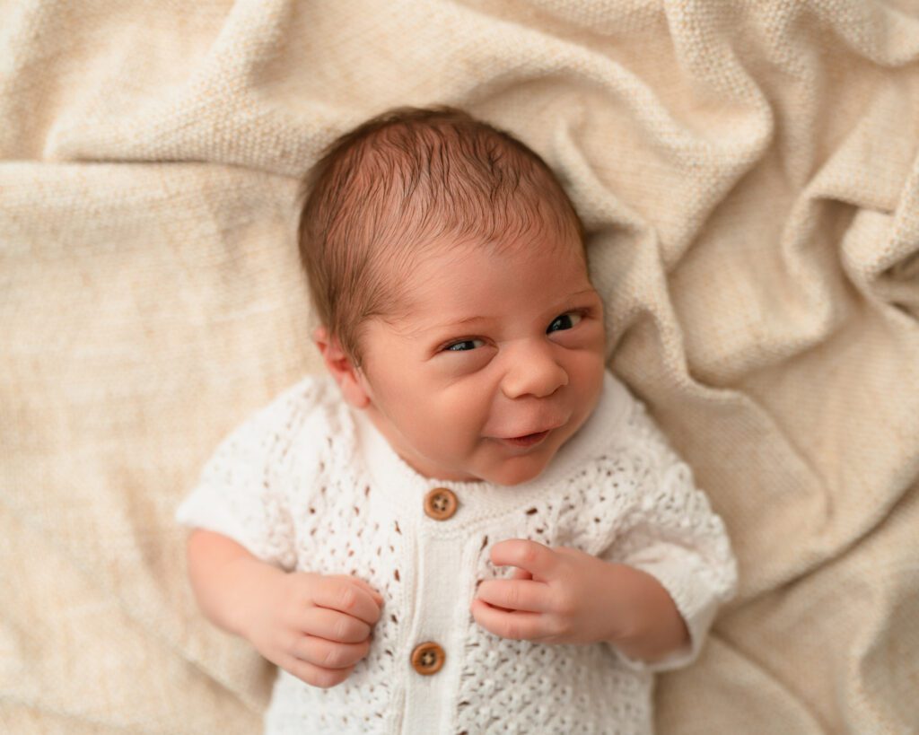 Newborn baby making a curious expression while lying on his back in a cream knit romper.