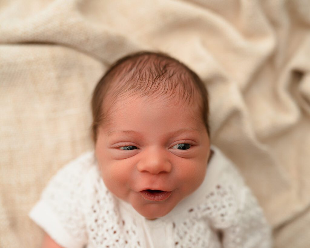 Awake newborn baby in a cream knit outfit looking toward the camera with a playful expression.
