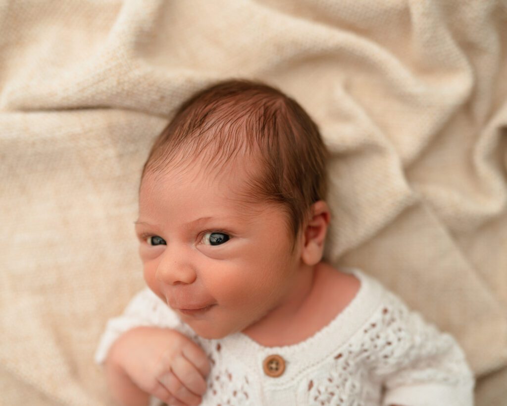 Awake newborn baby lying on his back with wide eyes and a playful expression on a neutral blanket.