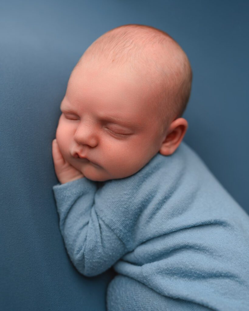 Baby boy in a soft blue romper resting his cheek on his hands against a matching blue backdrop.