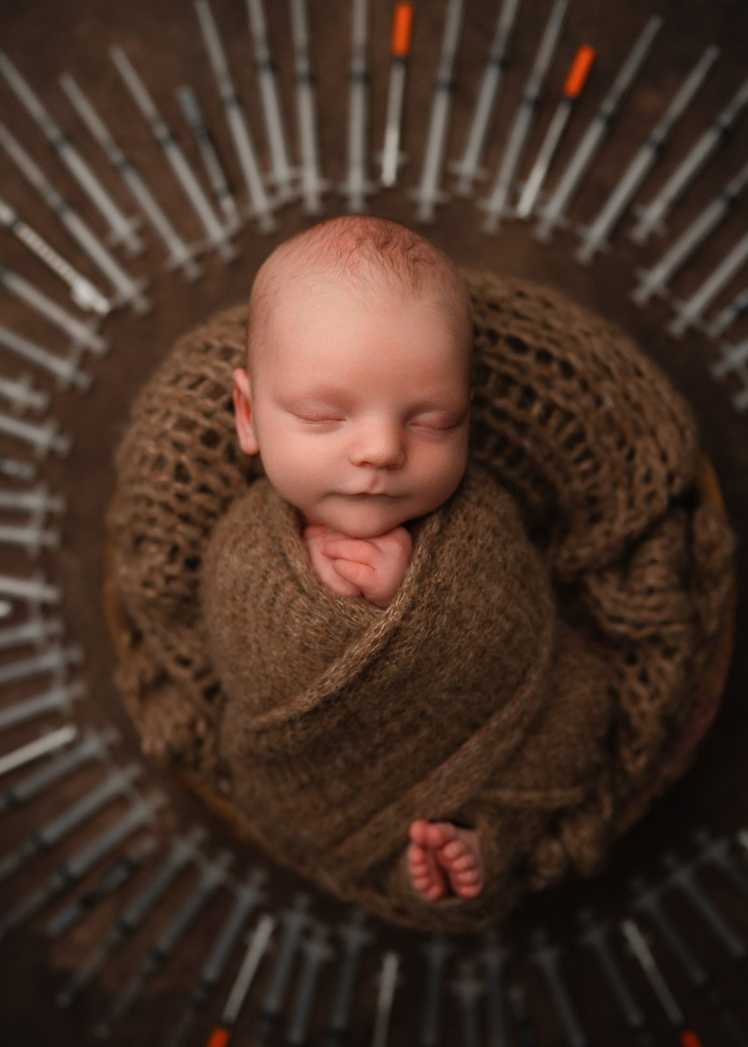 Sleeping newborn wrapped in brown knit surrounded by IVF needles for baby boy newborn photos in Atlanta.