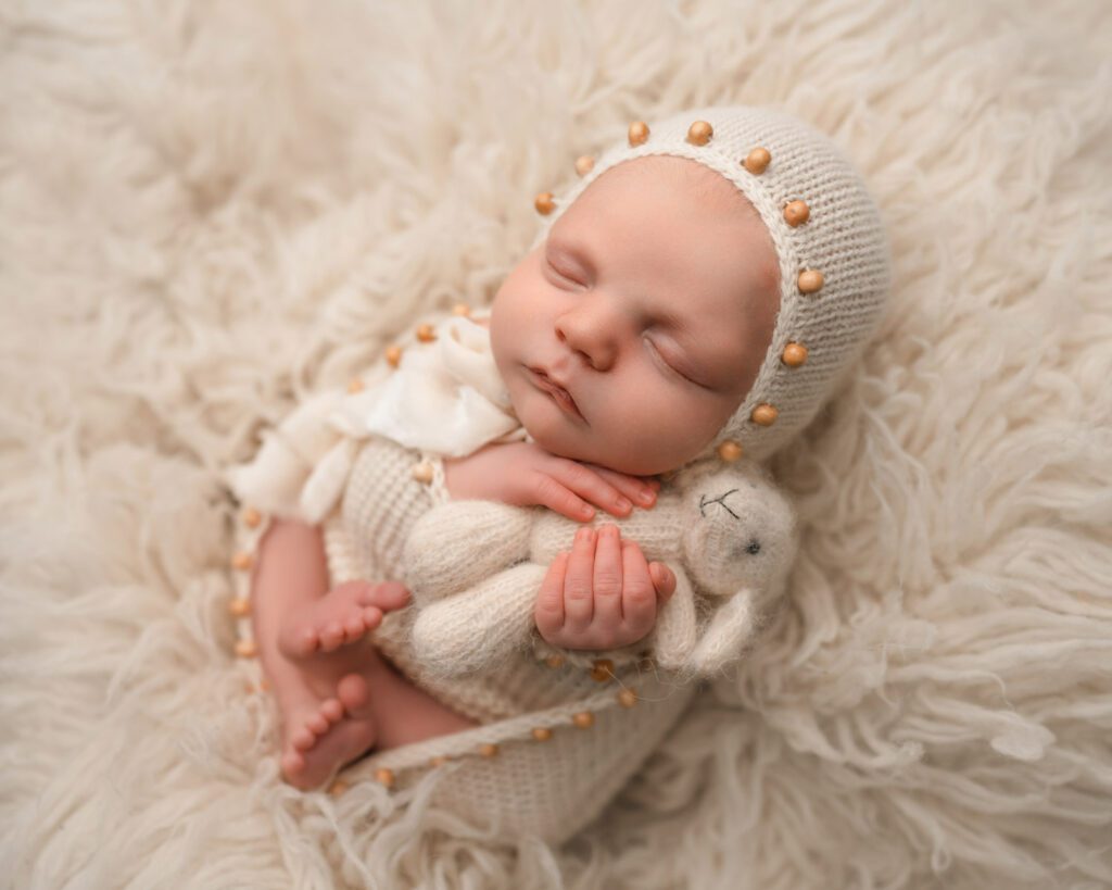 Baby boy holding a white bunny while wearing a cream knit bonnet with wooden bead accents on a fur blanket.