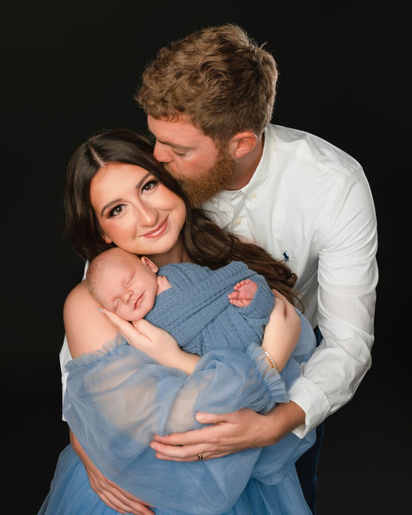 Dad kissing mom as she holds their sleeping newborn baby boy against a black studio backdrop.