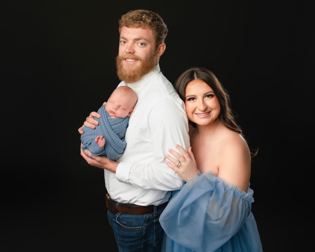 Mom in a blue tulle dress embracing dad as he holds their sleeping newborn baby boy.
