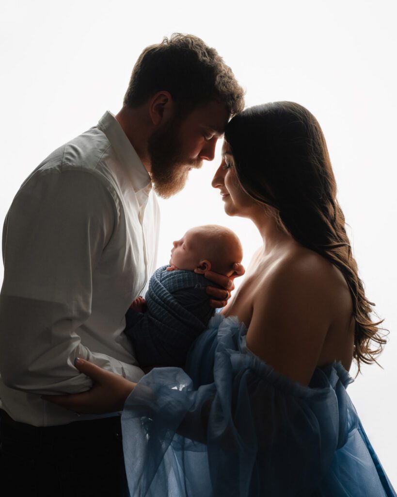 Parents touching foreheads holding their newborn baby boy between them with backlight lighting.