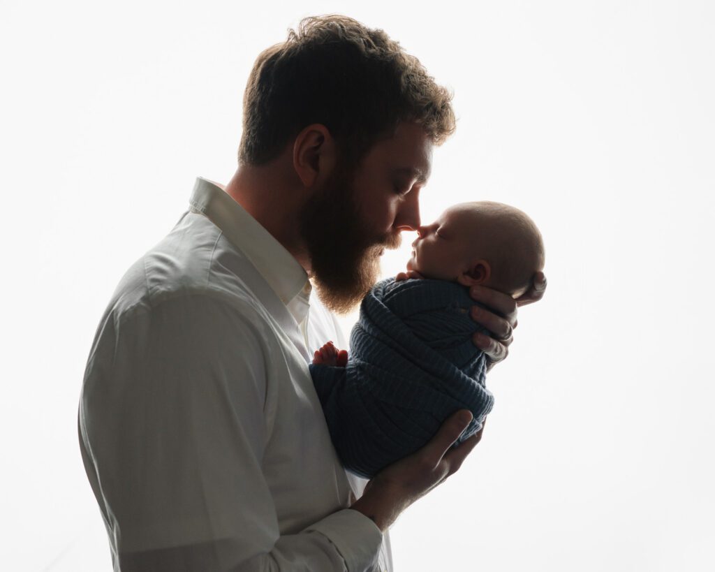 Dad holding his newborn son close as they touch noses against a bright white backdrop.