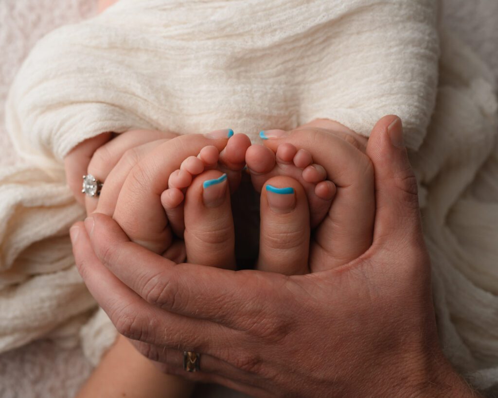 Parents holding their newborn baby boy’s tiny feet with mom’s blue nail polish visible.