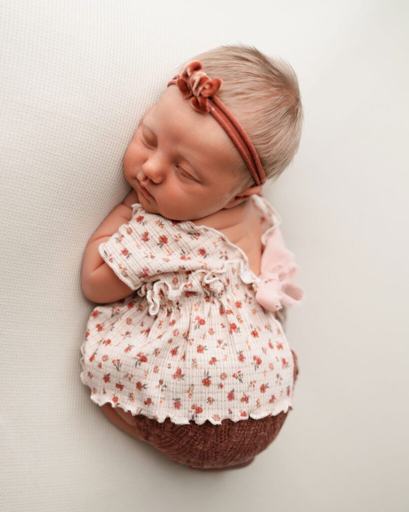 Overhead portrait of a sleeping infant in a pink floral outfit, maroon bloomers, and rust bow headband.