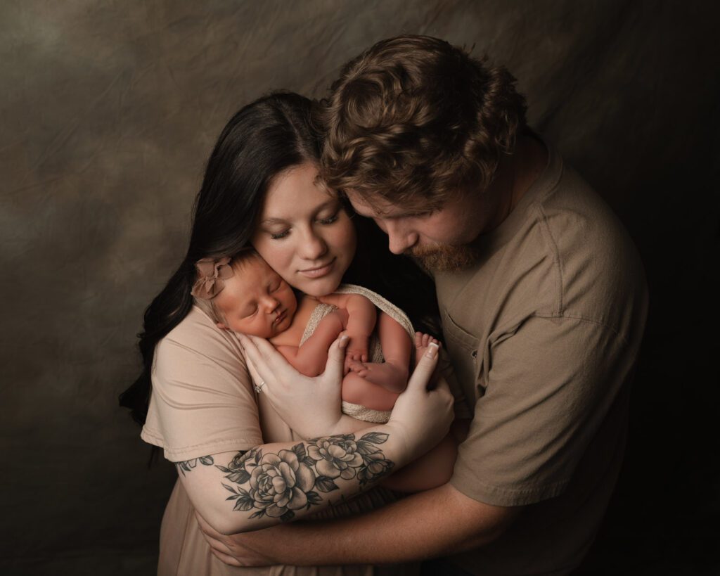 Mom and dad holding their newborn baby girl in neutral tan outfits against a warm brown studio backdrop.