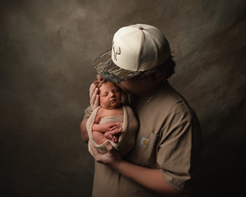 Dad holding his newborn daughter close and kissing her head as they wear matching tan tones against a brown backdrop.