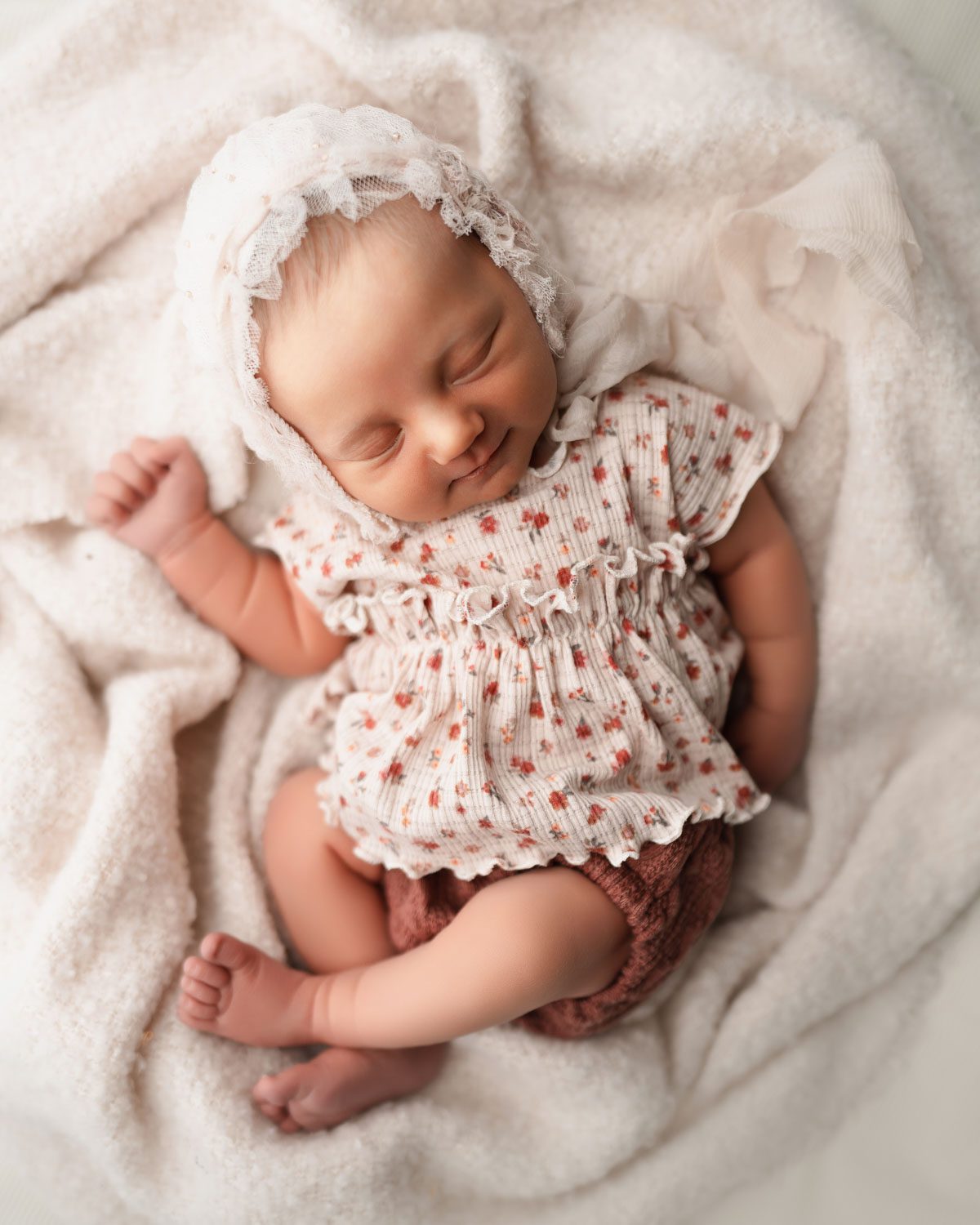 newborn girl in floral oufit and ruffled bonnet laying on soft cream fabric during atlanta baby girl newborn session