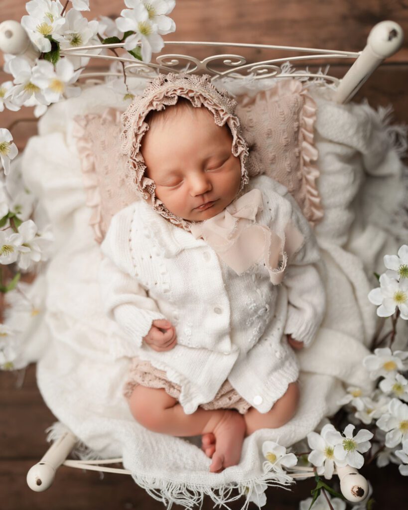 Sleeping newborn baby girl in a white knit sweater and peach ruffled bonnet on a flower-trimmed vintage bed.