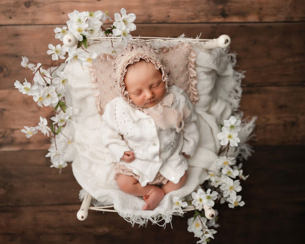 Sleeping newborn baby girl in a bonnet on a white bed surrounded by white flowers over hardwood floors.