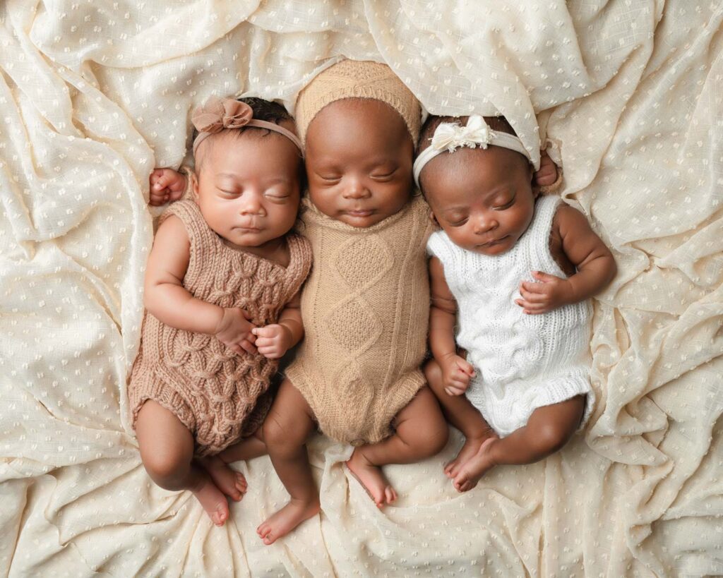 three newborn triplets sleeping together on soft textured fabric in cream, tan, and brown knit outfits.