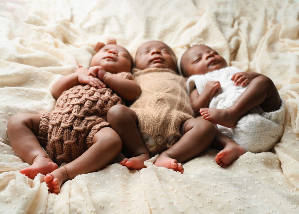 Three sleeping newborn triplets in coordinating tan, brown, and white knit outfits on a cream blanket.